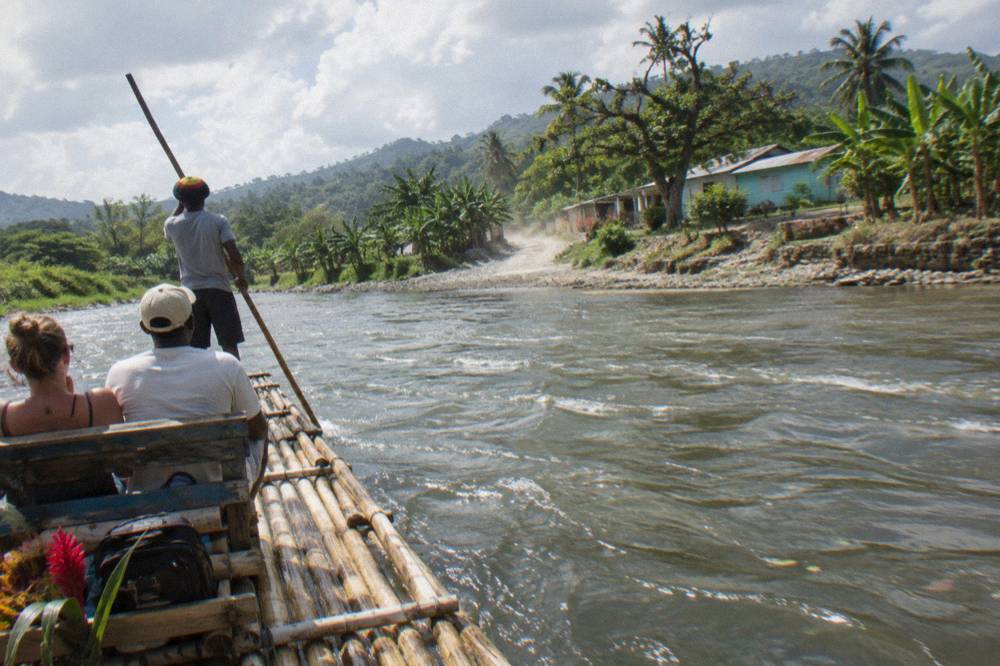 Wide bamboo raft floating on a slow river between tall trees, captain poling at the rear