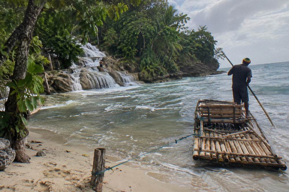 A single bamboo raft near the sand where a waterfall meets the sea, captain standing aboard with his pole