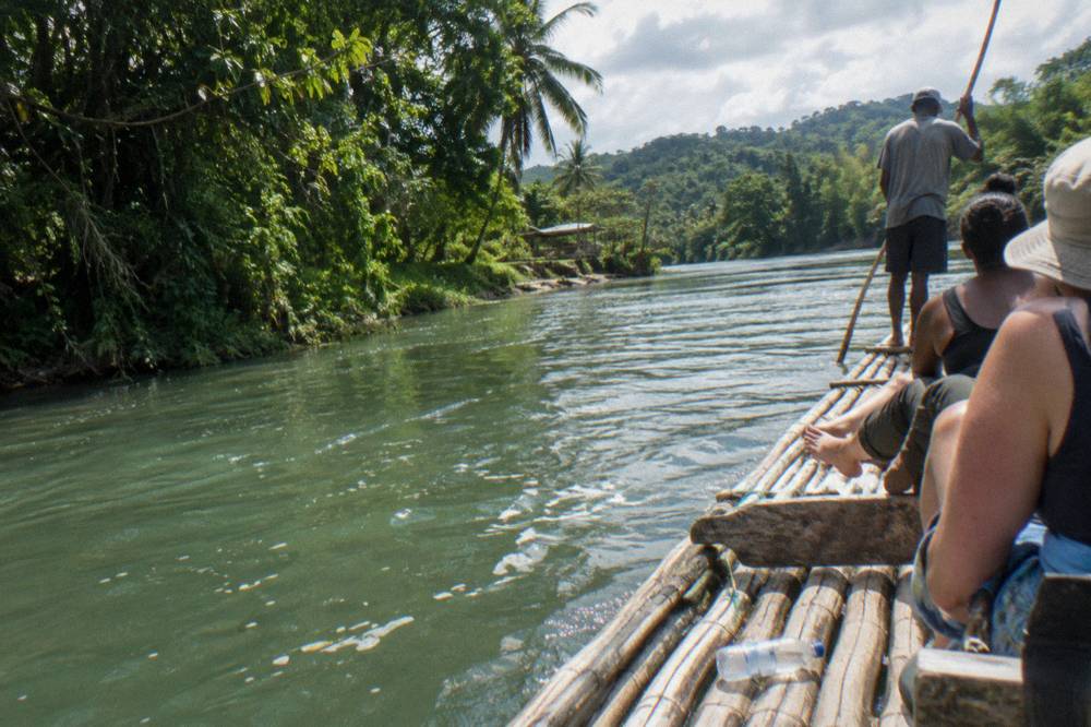 A bamboo raft captain standing at the rear of a raft on calm water between dense trees