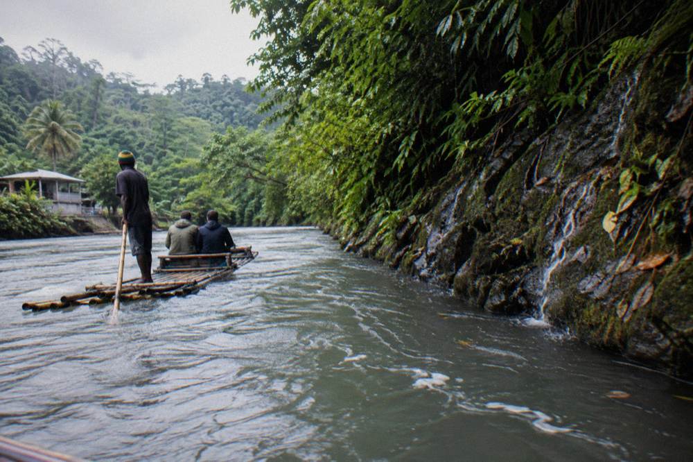 Bamboo raft midstream on the Great River in grey light, captain poling, two passengers aboard