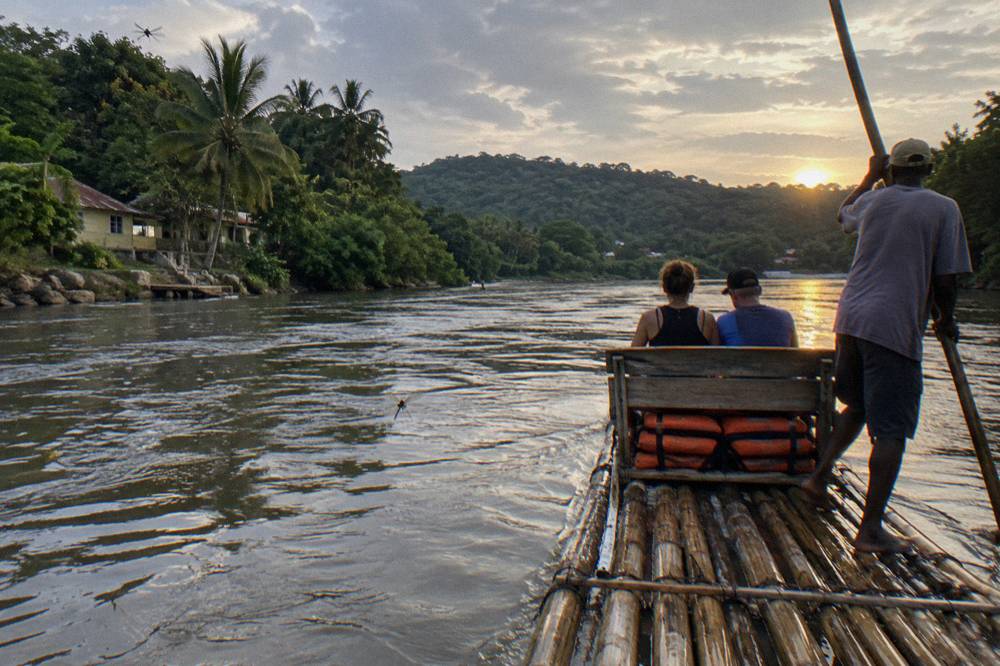 A raft captain standing at the rear of a bamboo raft poling along a calm river