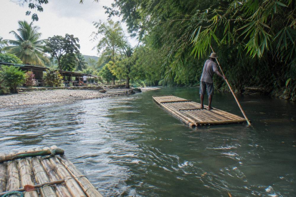 A raft captain poling an empty bamboo raft along a quiet green river corridor