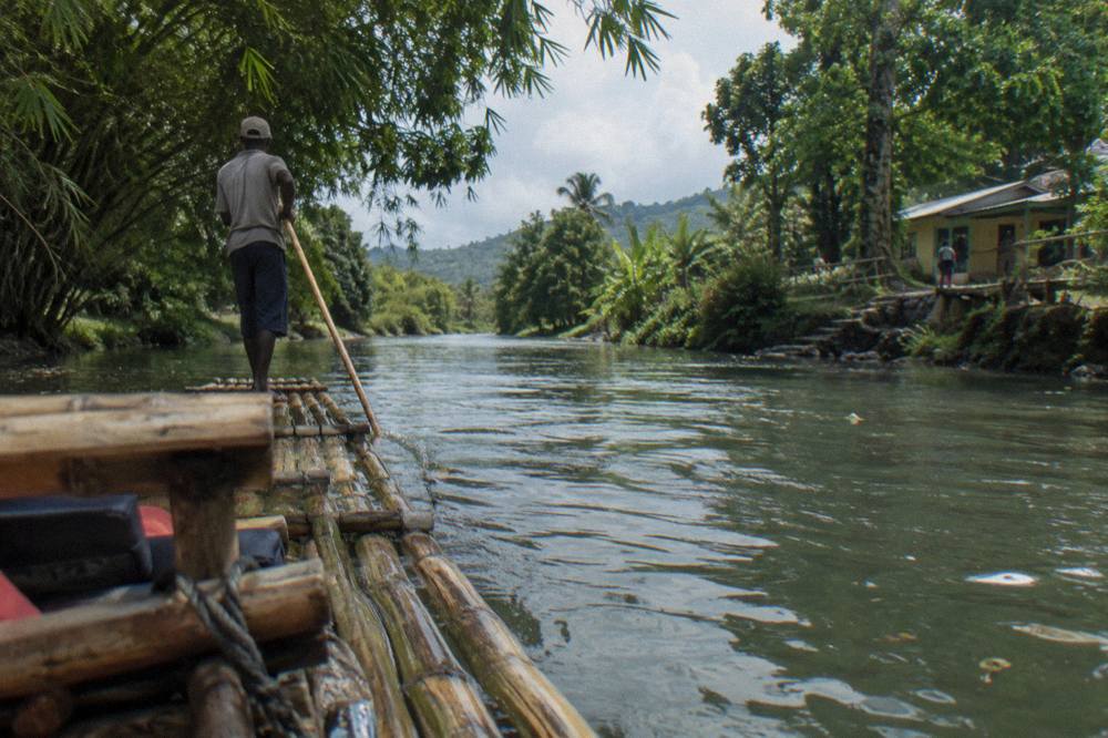 A raft captain standing at the rear of a bamboo raft poling it along a slow green river between overhanging trees