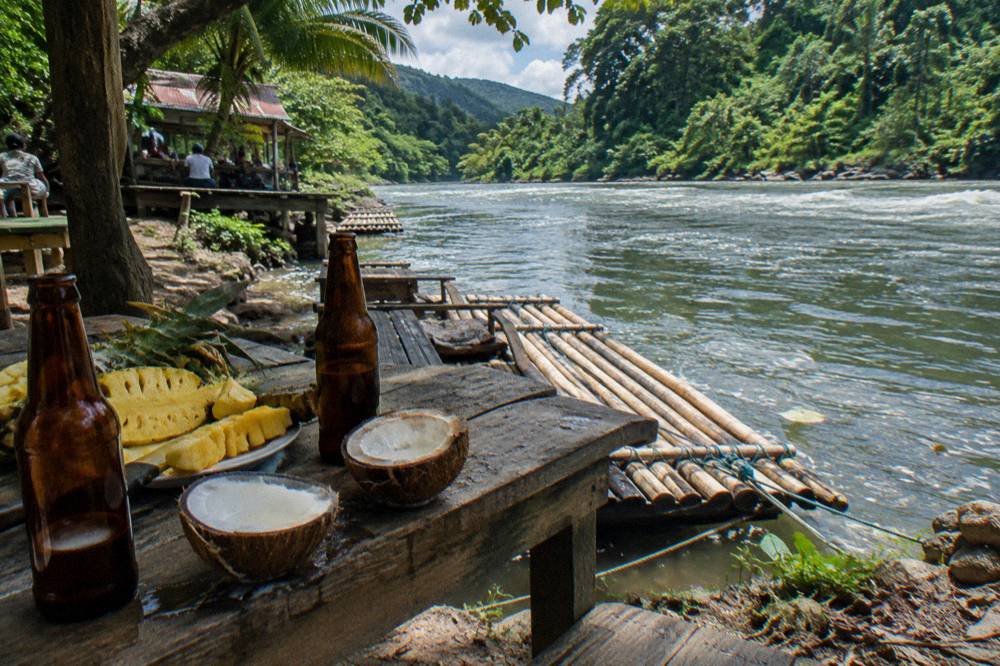 A shaded table on a riverbank holding fresh fruit, coconut and cold drinks beside a moored bamboo raft