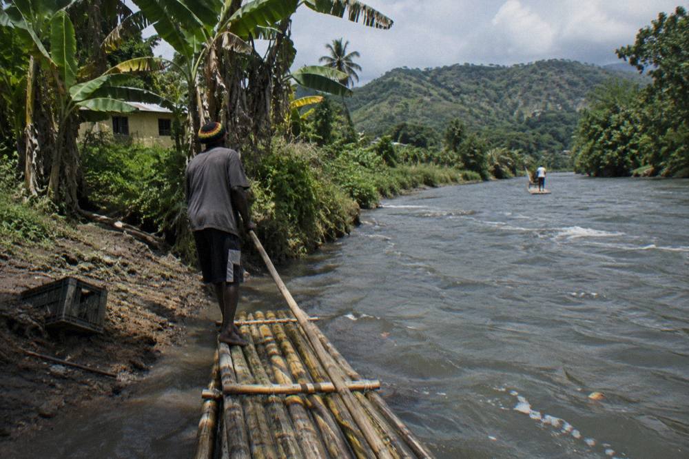 A bamboo raft resting on a riverbank beside tall banana trees and green hills