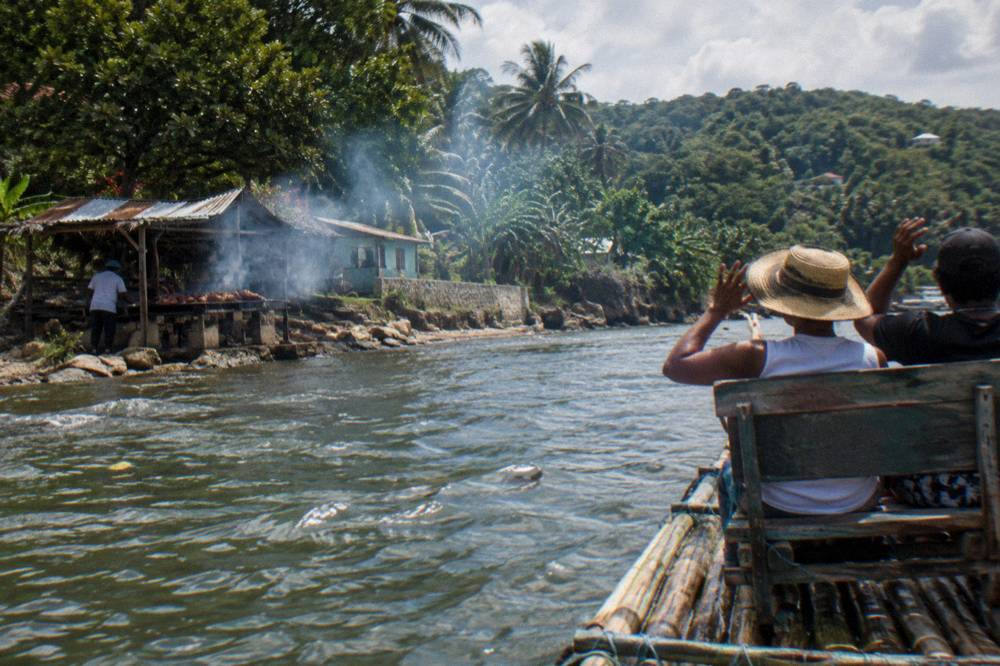 Bamboo raft gliding on a river near Port Antonio, two passengers on the bench, smoke drifting from the riverbank