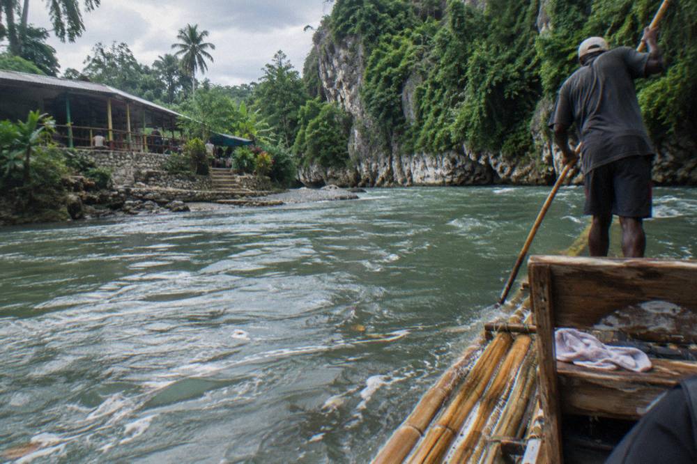 A captain standing at the rear of a bamboo raft, poling along a wide green river below limestone cliffs