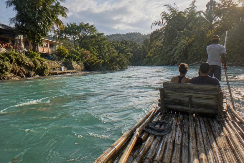 Two guests seated on a bamboo raft as the captain poles it along a wide river past a forested bank
