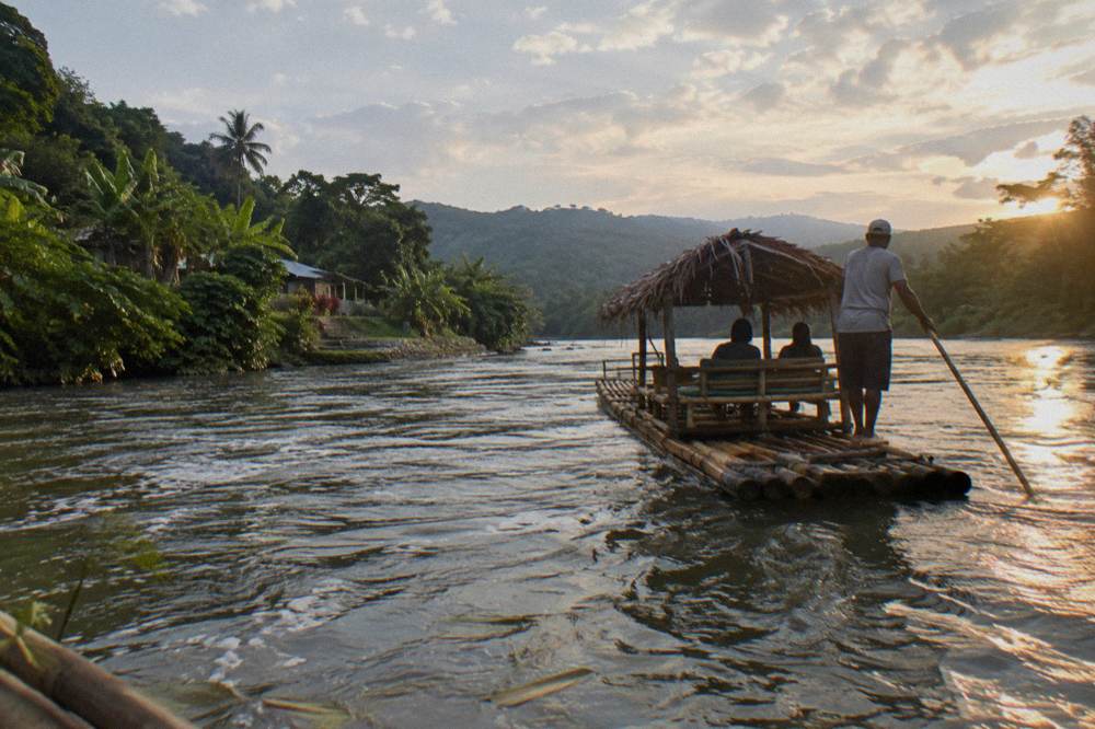 A long bamboo raft with a thatched seat gliding on calm water between forested banks