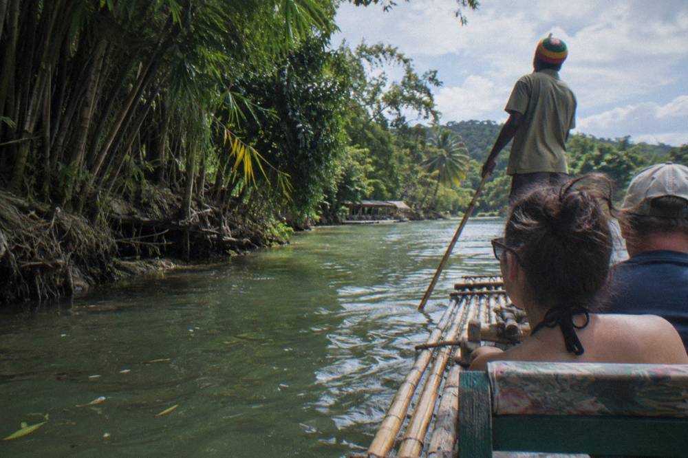 A raft captain standing at the rear of a bamboo raft guiding it through still green water