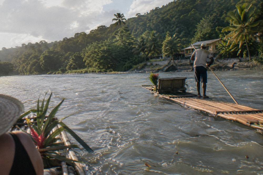 A long bamboo raft poled by a captain drifting down a wide green river between forested riverbanks