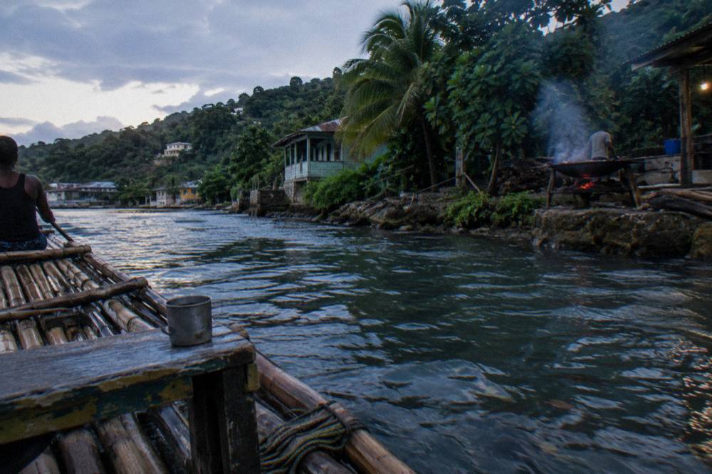 A bamboo raft drifting a calm river at dusk, pimento wood smoke rising from a jerk pan on the bank