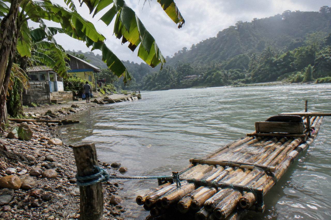 A bamboo raft resting on the banks of a wide green river beneath forested hills in Portland, Jamaica