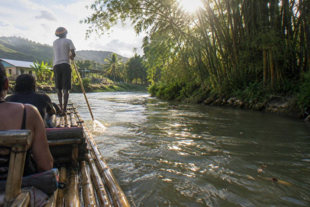 A bamboo raft drifting past tall bamboo stands along a narrow green riverbank