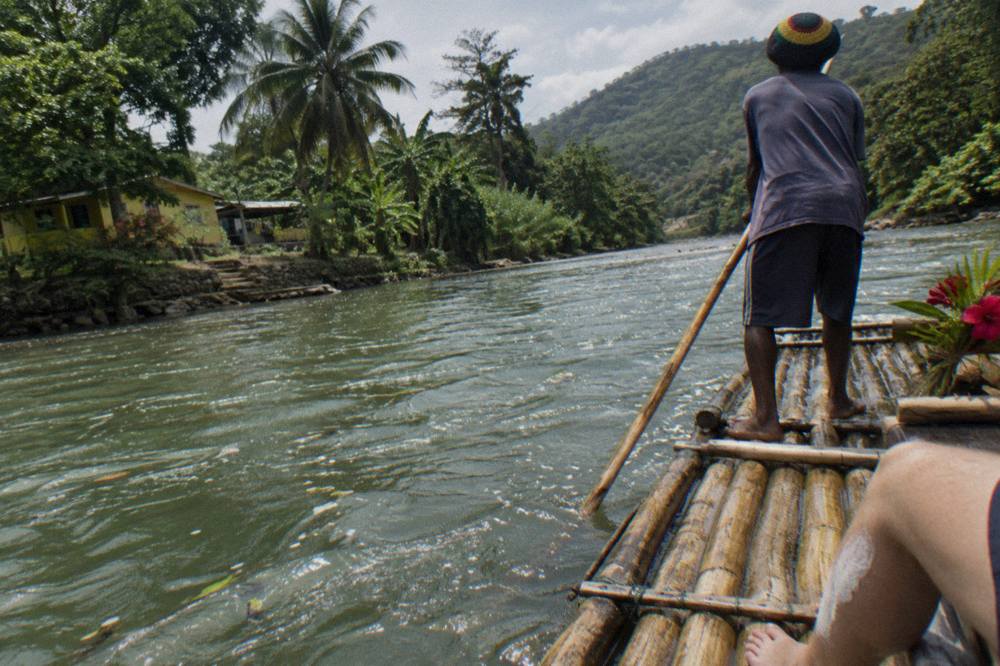 A raft captain rubbing limestone paste on a seated passenger's leg as the raft drifts along a green river