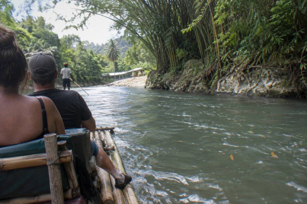 Two passengers seated on a bamboo raft beside tall bamboo and greenery along a slow river bend