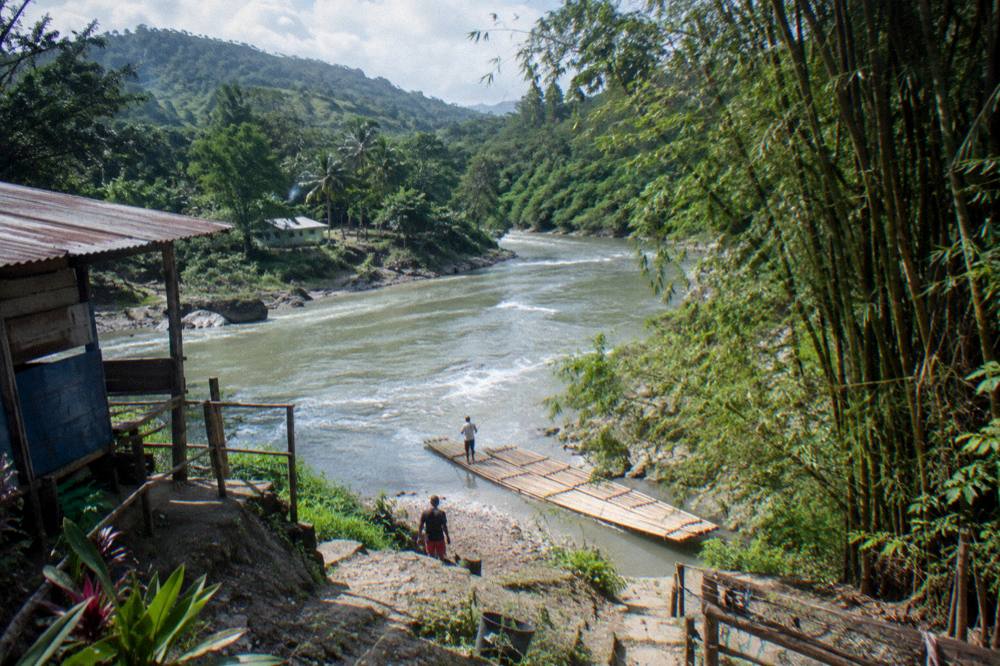 A river winding through green hills toward a small rafting village with bamboo stands on the bank