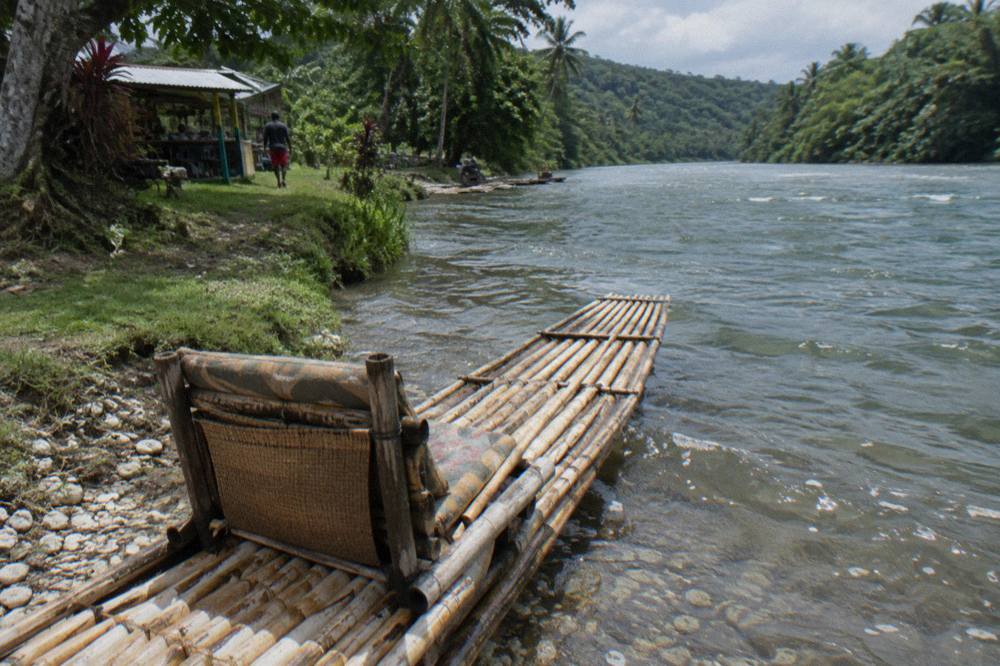 A long bamboo raft with a woven padded seat resting at a calm riverbank launch point