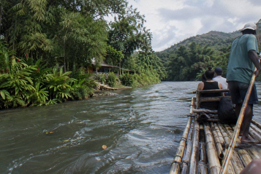 A single bamboo raft gliding slowly past a green forested riverbank in soft light
