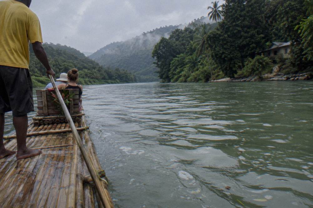 A long bamboo raft floating on wide calm water between tree-lined banks in warm light