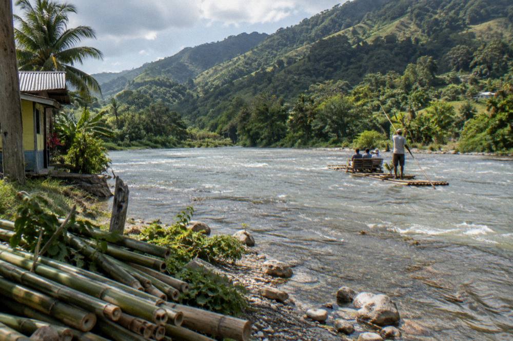 Sunlight returning over a calm river valley with a bamboo raft poling downstream in the distance