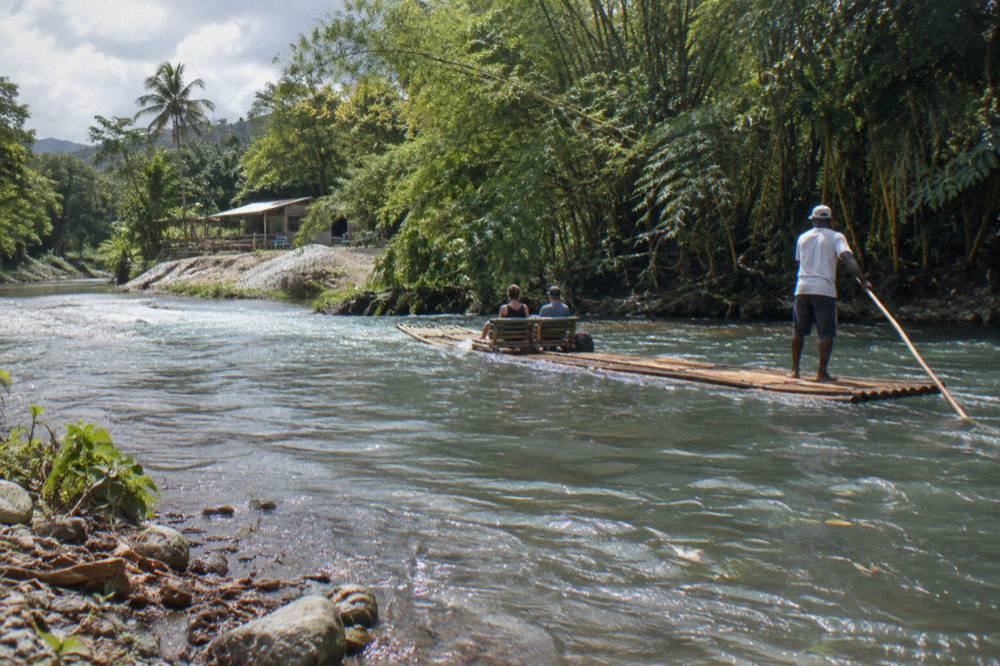 A raft captain standing at the rear of a bamboo raft poling along a calm green river