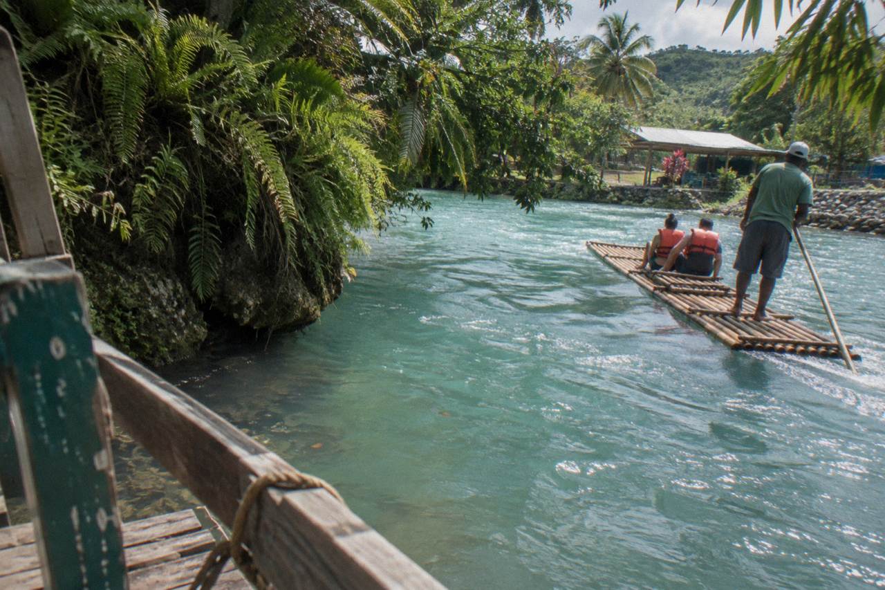 A long bamboo raft floating calmly on a wide green Jamaican river between tree-lined banks