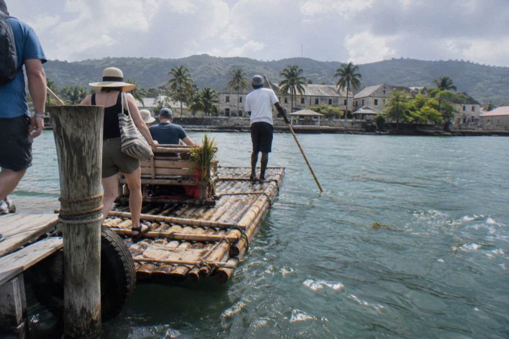 A captain poling a bamboo raft down a calm river between green hills