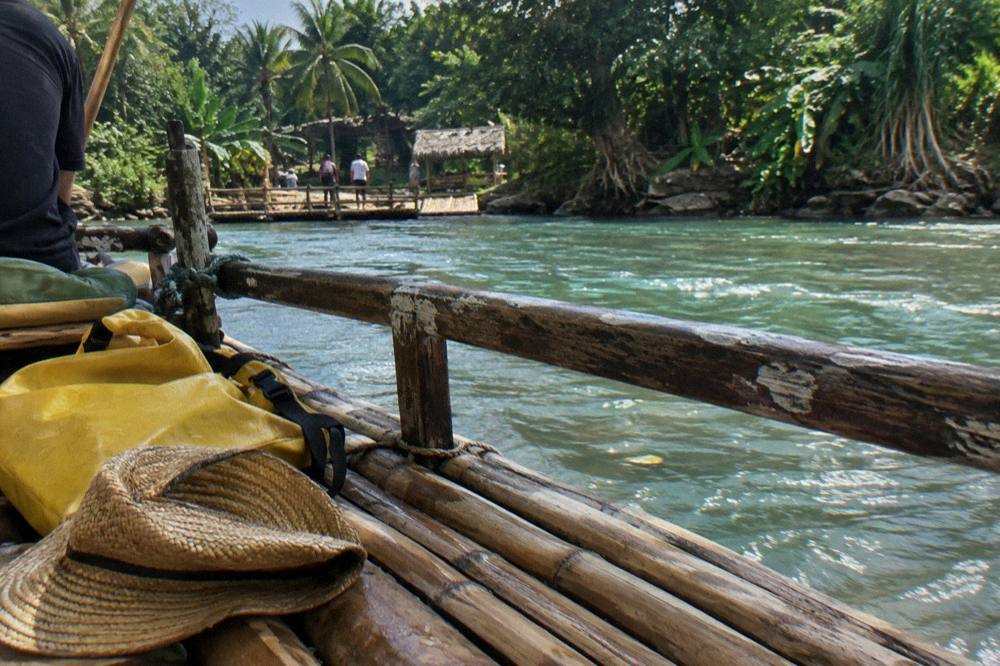 A folded straw hat and dry bag resting on a wooden bench beside a calm river