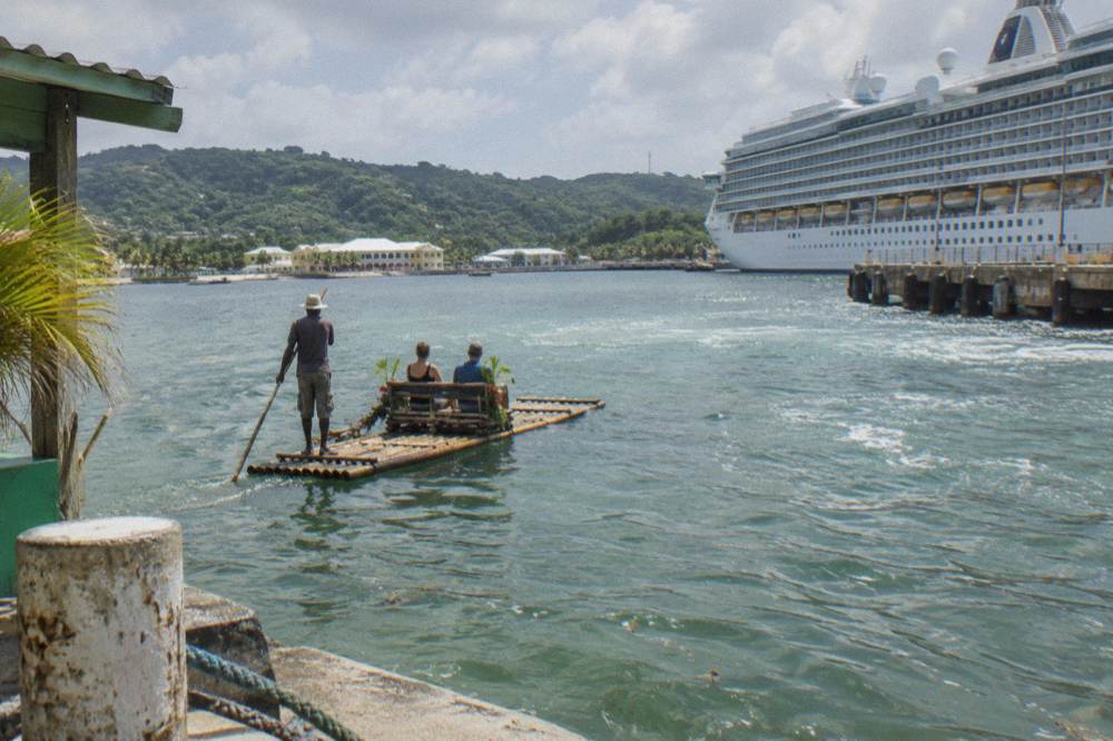 A bamboo raft with two passengers and a poling captain on calm green water below wooded hills