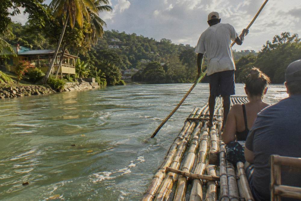 A raft captain standing at the rear of a bamboo raft poling along a calm river