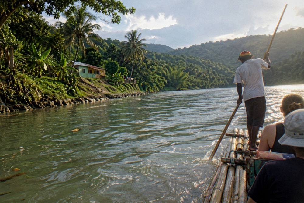 A captain poling a bamboo raft along a wide green river toward forested hills