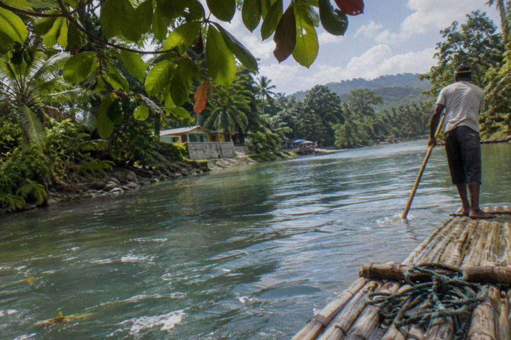 A bamboo raft drifting on calm turquoise water between green riverbank trees