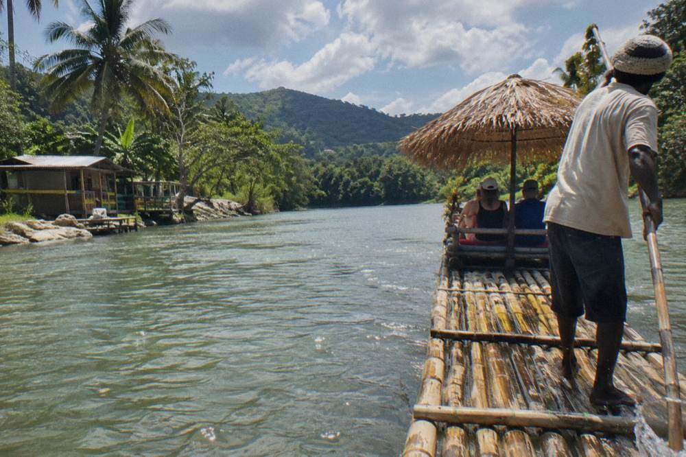 A raft captain standing at the rear of a bamboo raft poling along a wide calm river