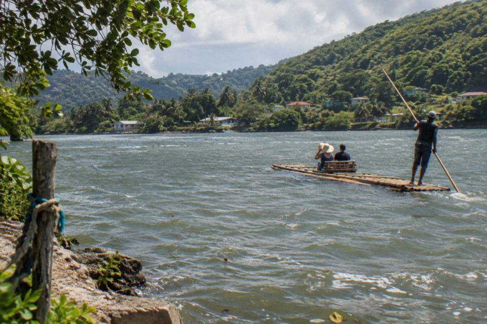 A captain poling a long bamboo raft along a wide green river between forested hills