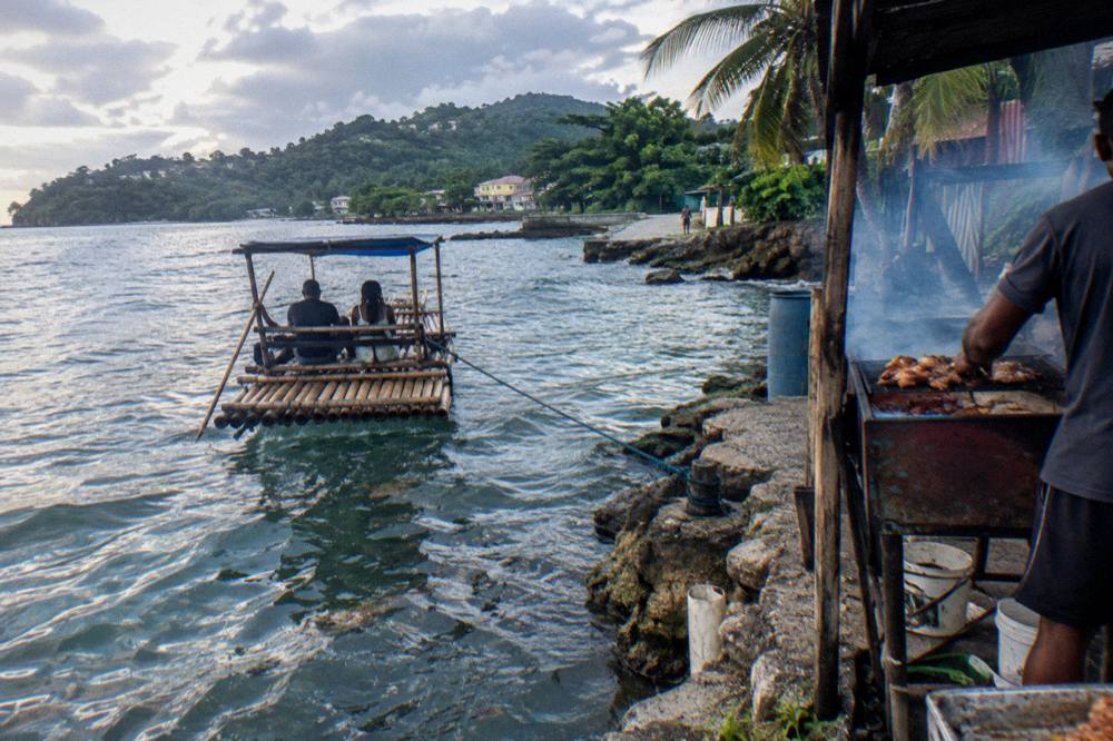 Bamboo raft with two passengers waiting as jerk smoke rises from a roadside grill holding chicken halves