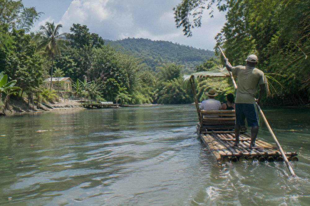 A raft captain standing at the rear of a bamboo raft with a long pole on quiet water
