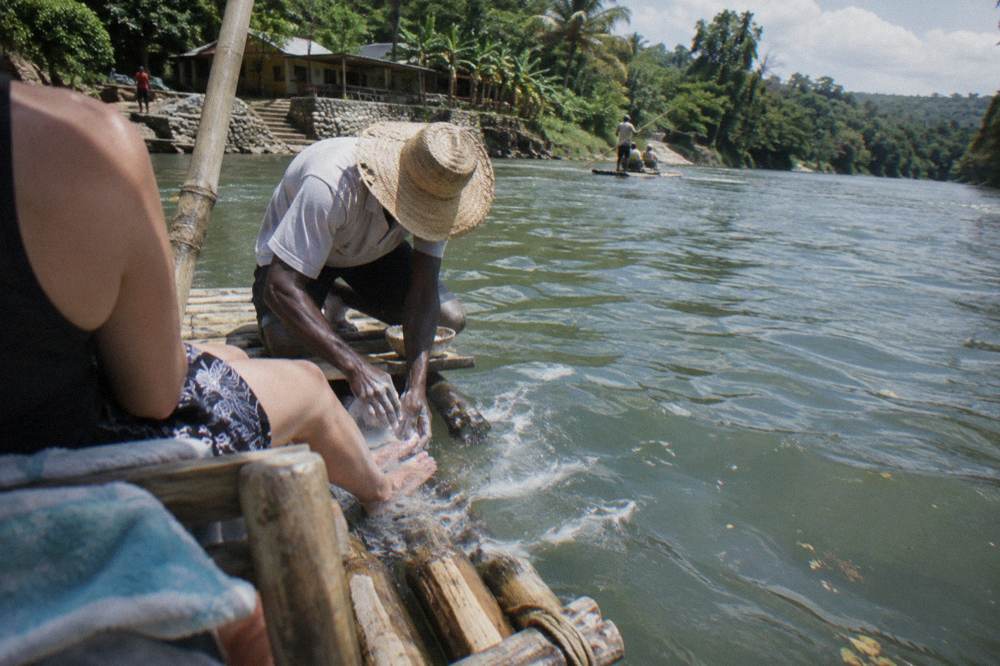 A captain kneeling at the front of a bamboo raft performing a limestone foot massage in shallow water