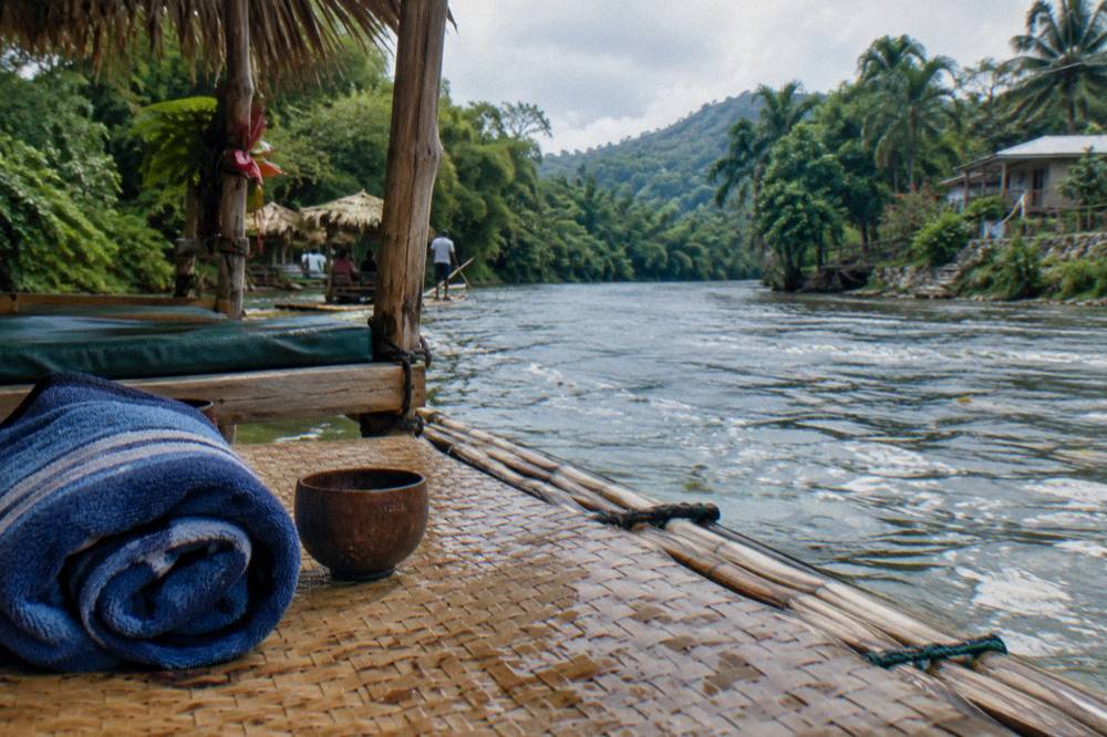 Woven bamboo mat on a moored raft deck with a rolled towel, a wooden cup and rain-spots on the weave