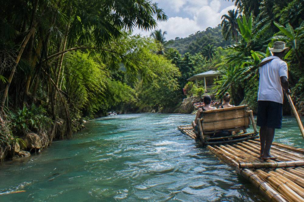 A long bamboo raft with the captain standing and poling, two passengers on the woven bench, bamboo banks closing over turquoise-green water