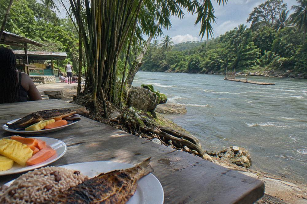 A spread of grilled fish, rice and tropical fruit on a wooden table beside a river